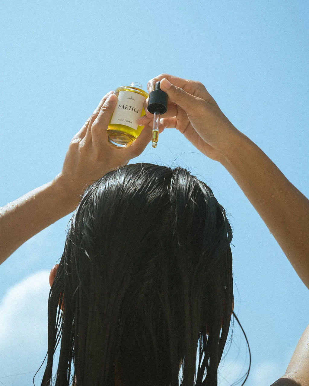 Woman applying hair oil to scalp against a clear blue sky