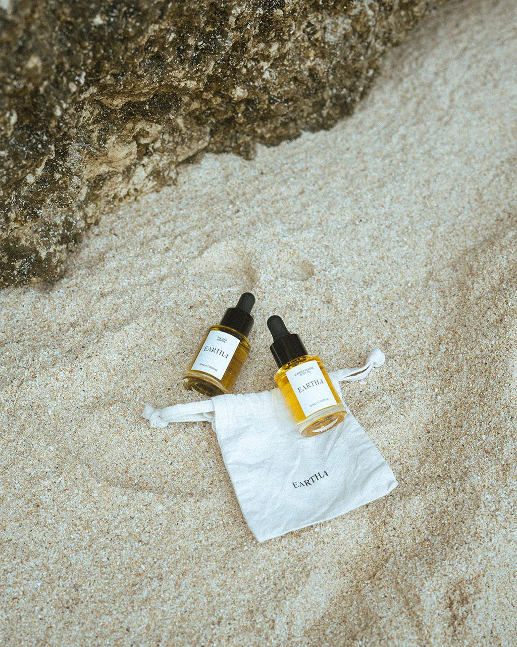 Two bottles of Eartha travel-sized body and hair oil with a brand pouch on sandy ground