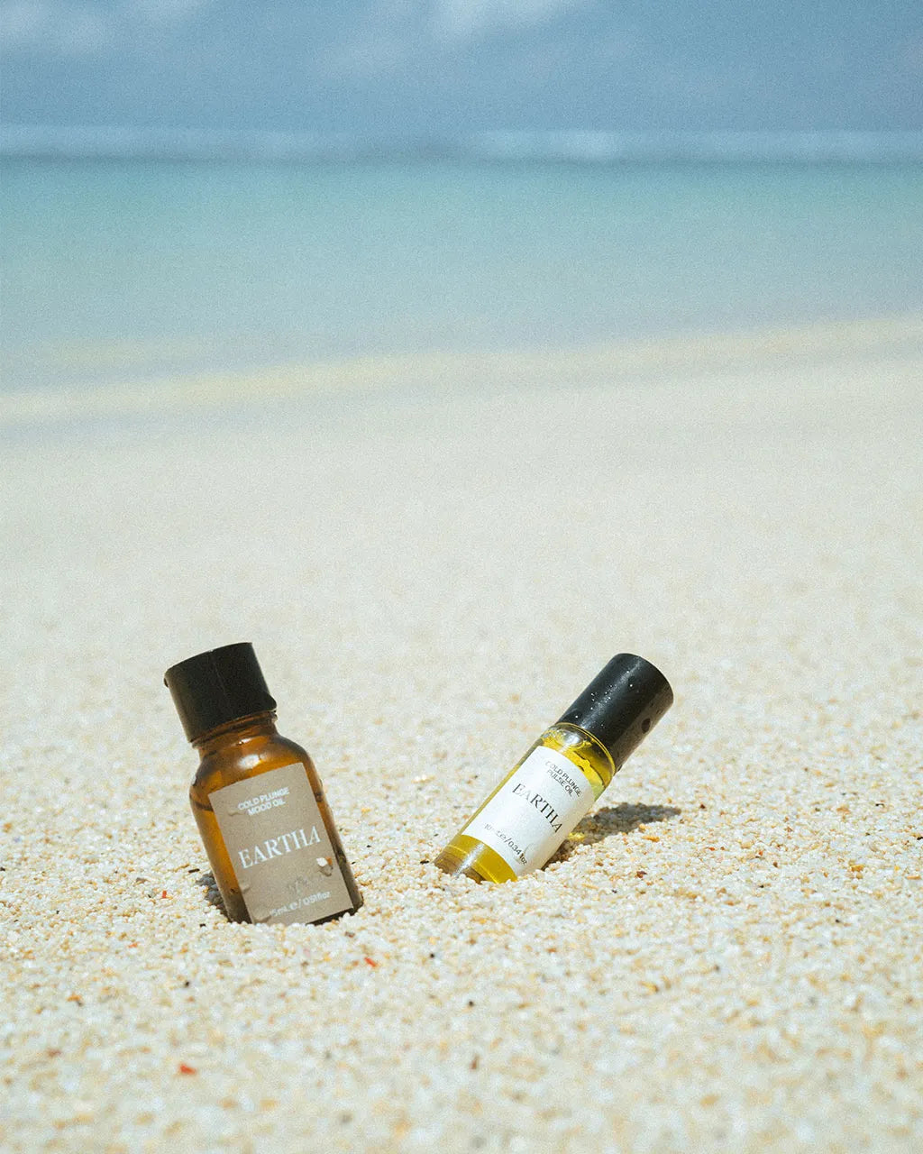 Two small bottles of essential oil and essential oil roll-on on a sandy beach with ocean and sky in the background.