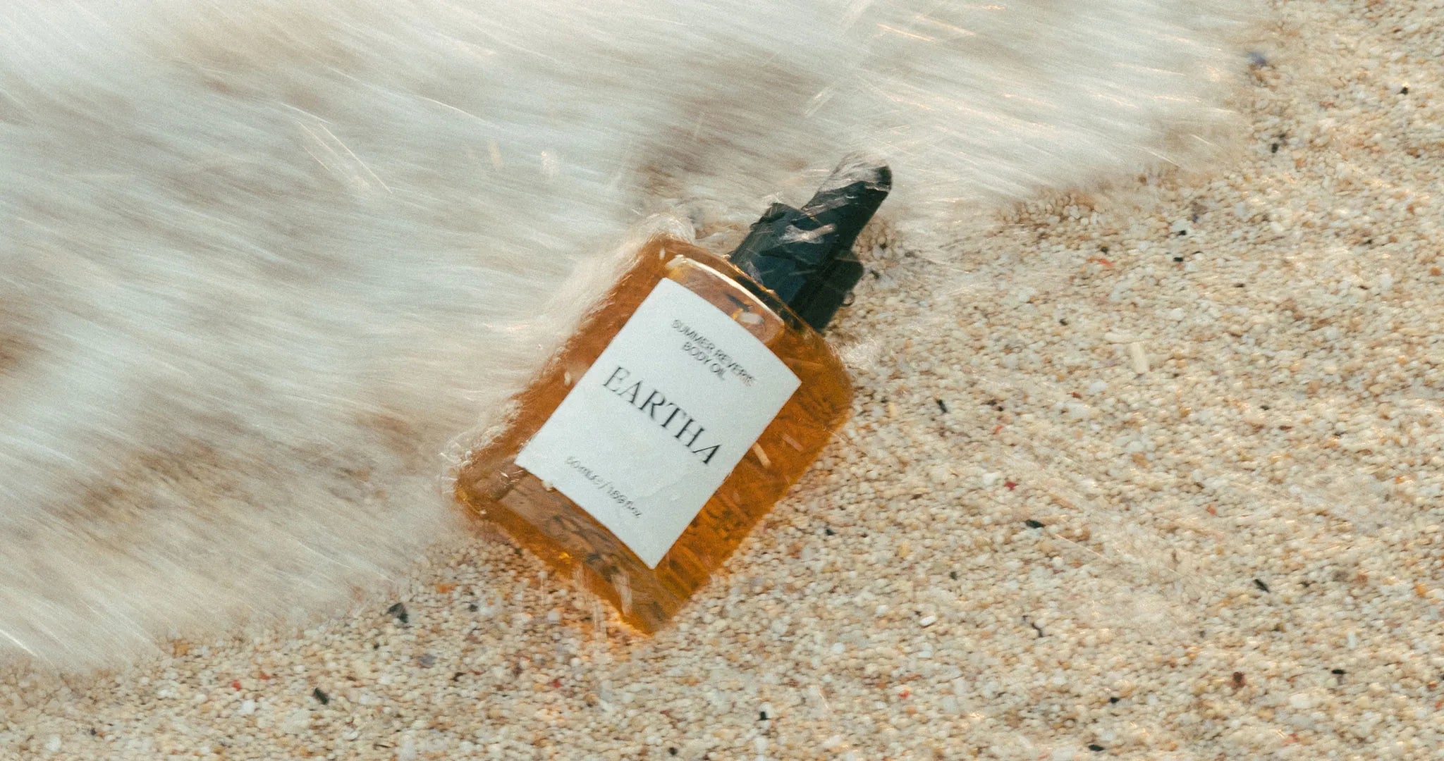 Bottle of Eartha Summer Reverie Body Oil on sand with water flowing over it