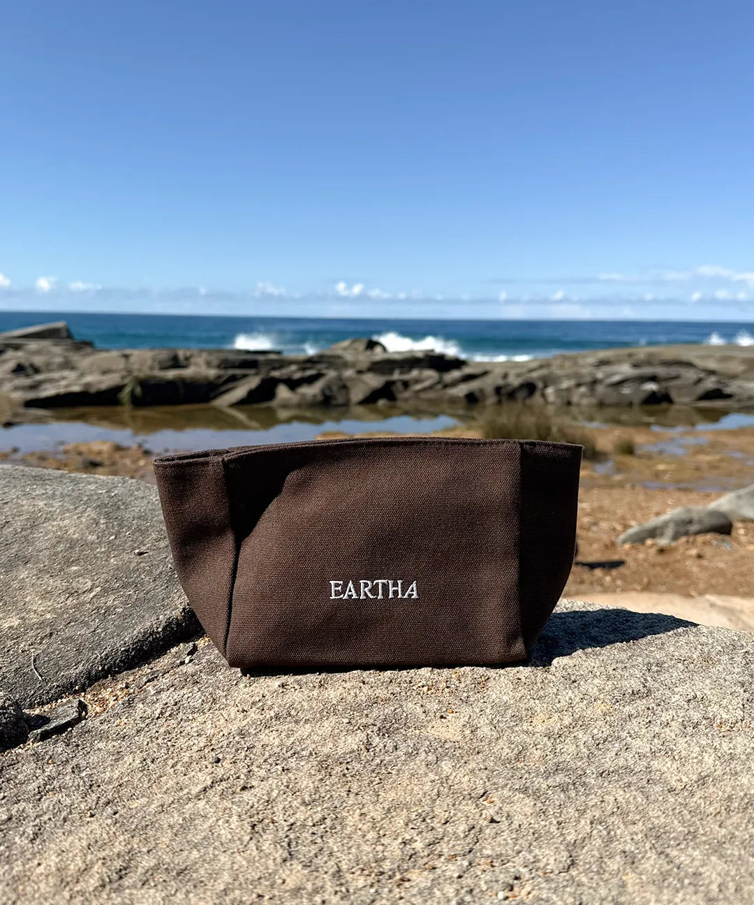 A dark brown canvas pouch with the brand name 'EARTHA' embroidered in white on the front, resting on a large rock with a rocky shoreline and blue ocean in the background.