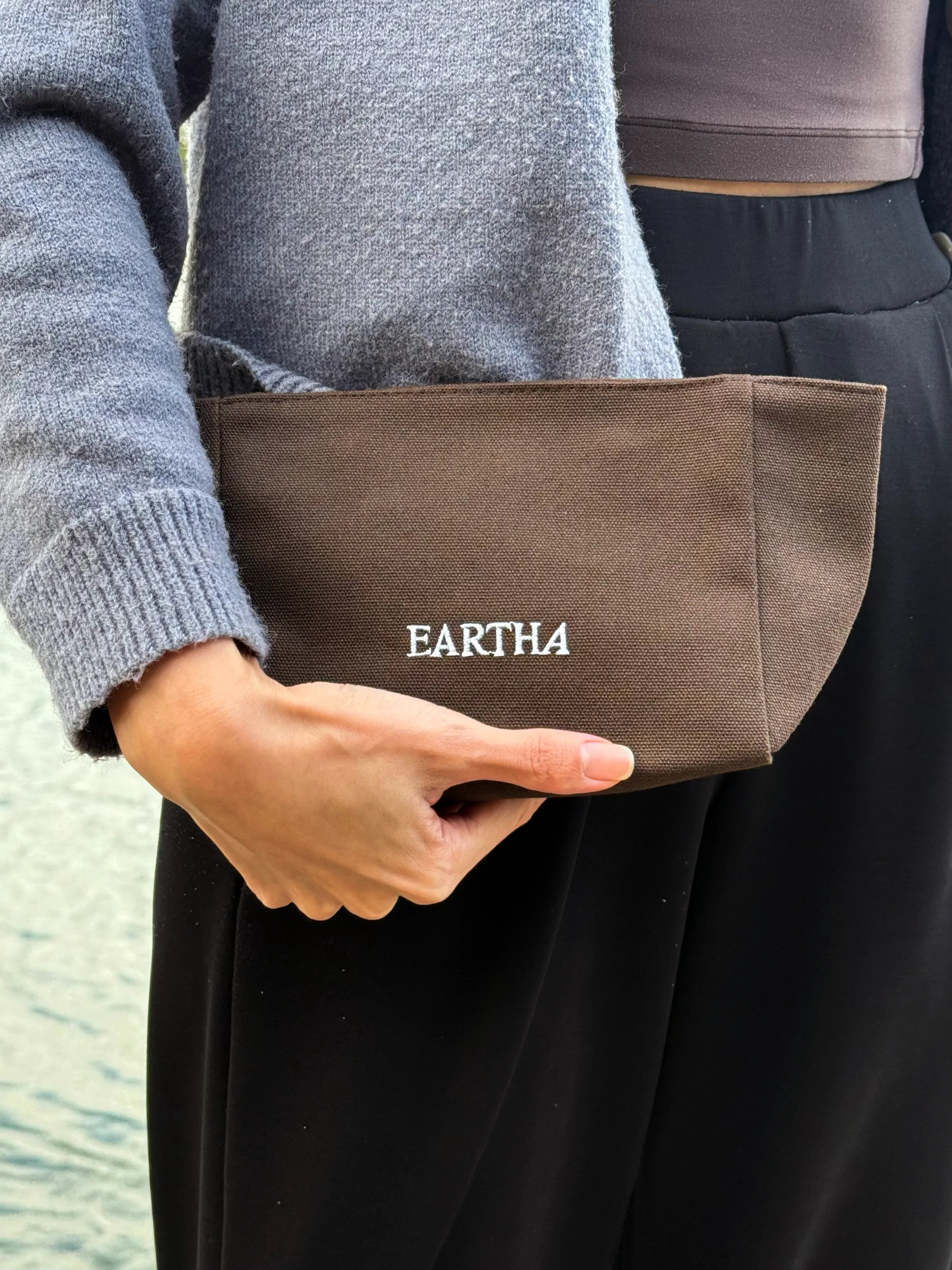 A person wearing a blue knit sweater and black pants holds a brown fabric pouch with the brand name 'EARTHA' embroidered in cream on the front.