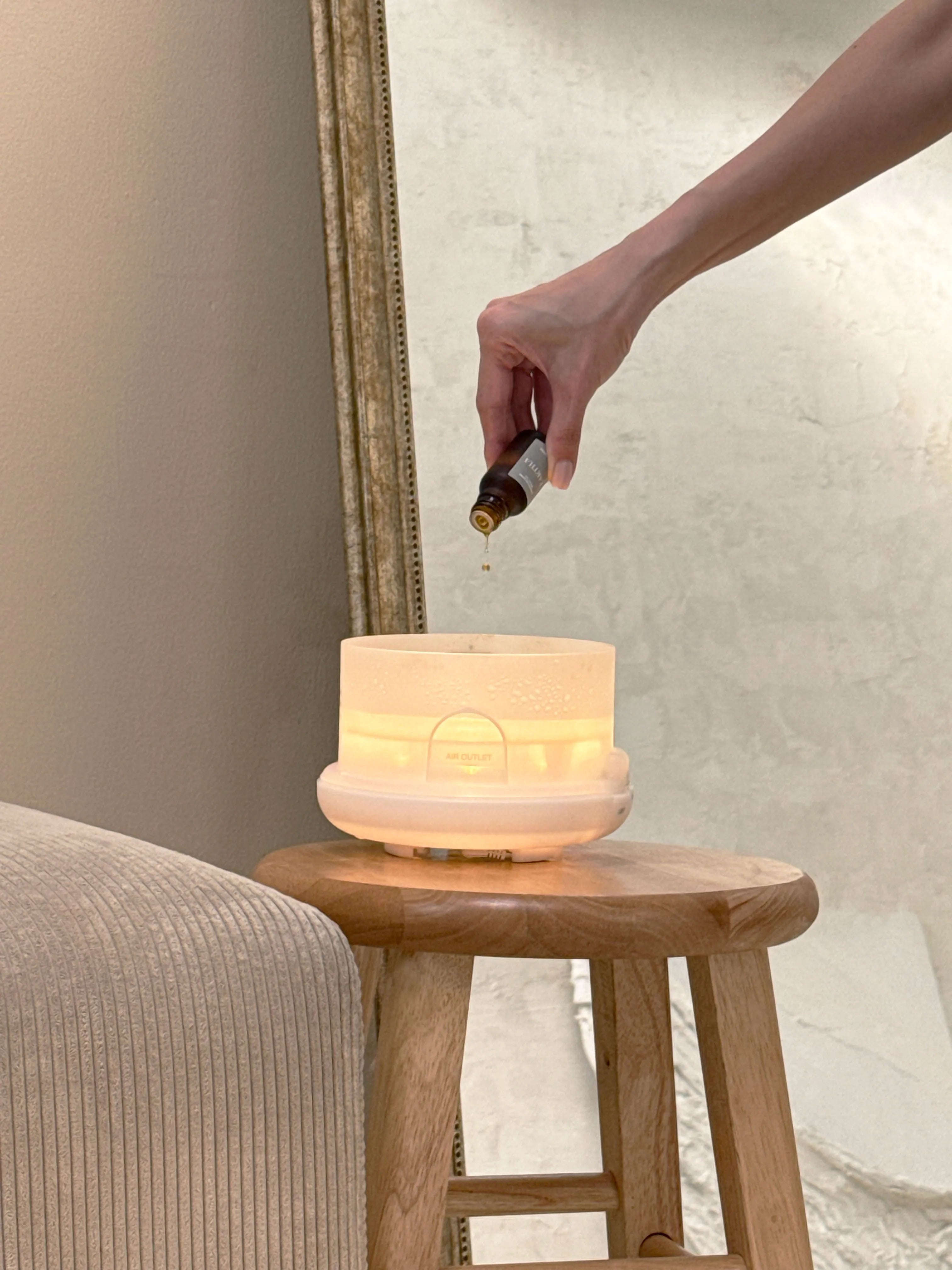 Hand pouring Eartha essential oil into a diffuser on a wooden stool with a neutral background