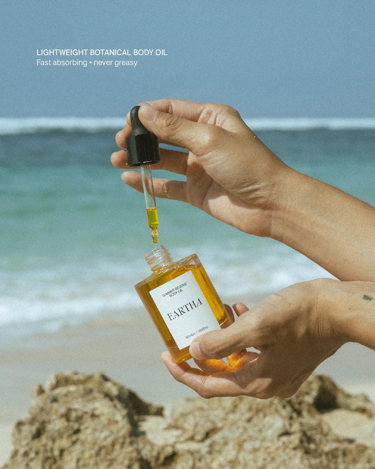 Person holding a bottle of Eartha oil with a dropper over a beach background