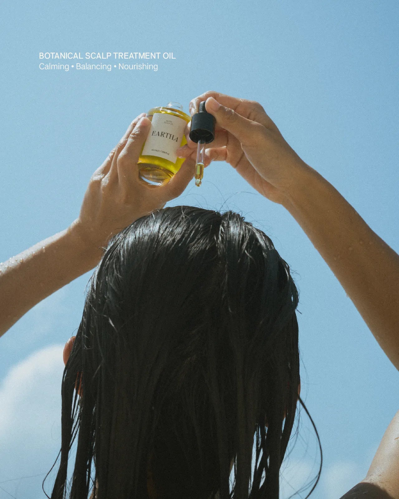 Woman applying a dropper of oil to their scalp against a clear blue sky