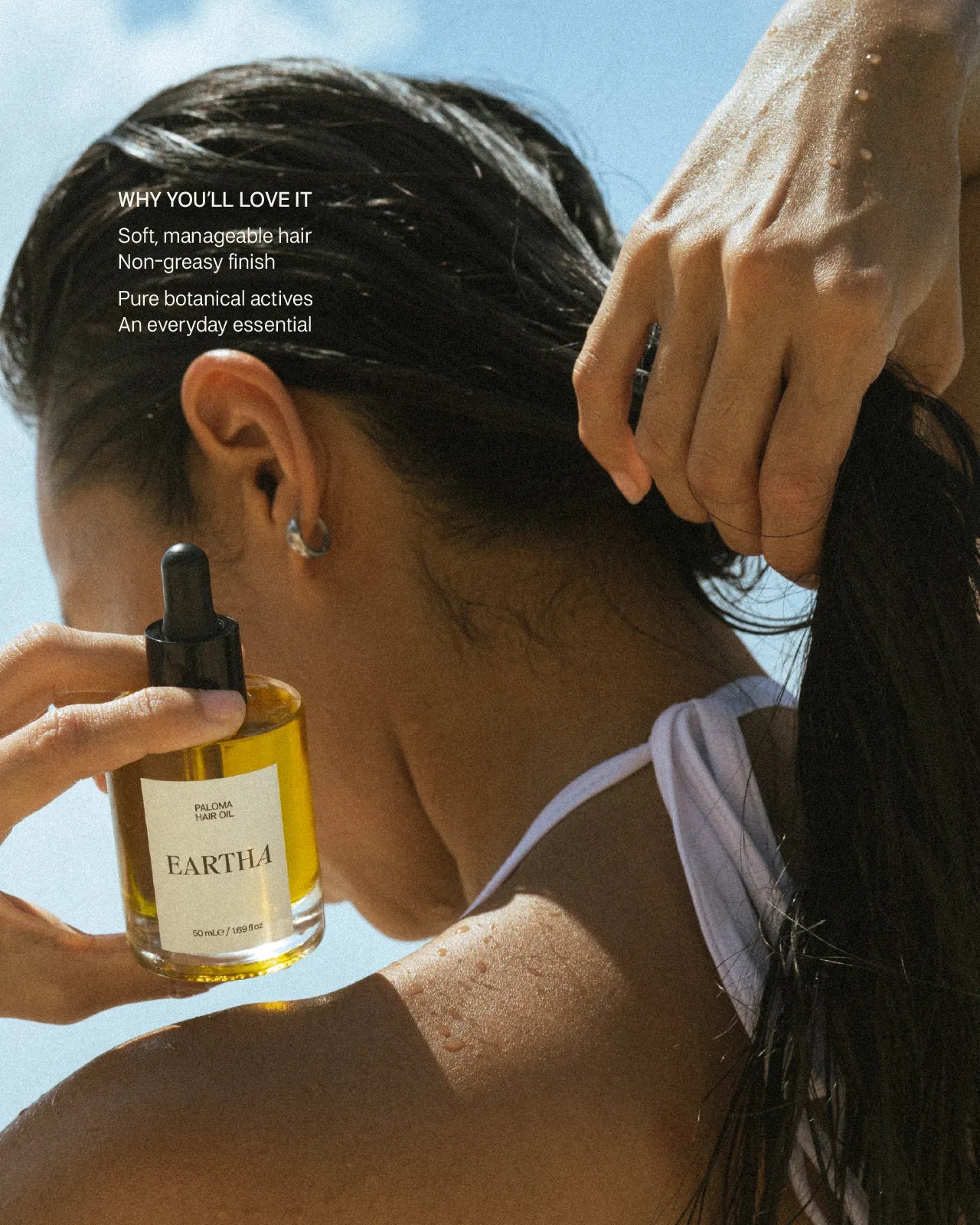 Woman holding a bottle of Eartha hair product with another hand touching their hair.