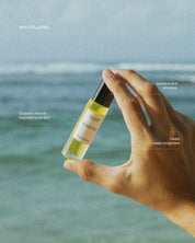 Hand holding a bottle of Eartha essential oil roll-on product with ocean background