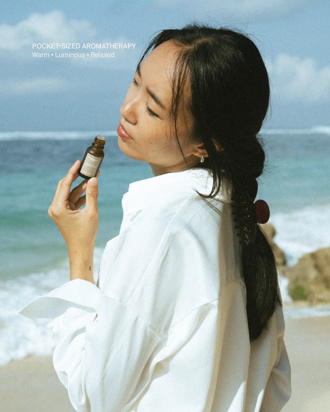 Woman holding a bottle of essential oil on a beach with ocean and sky in the background