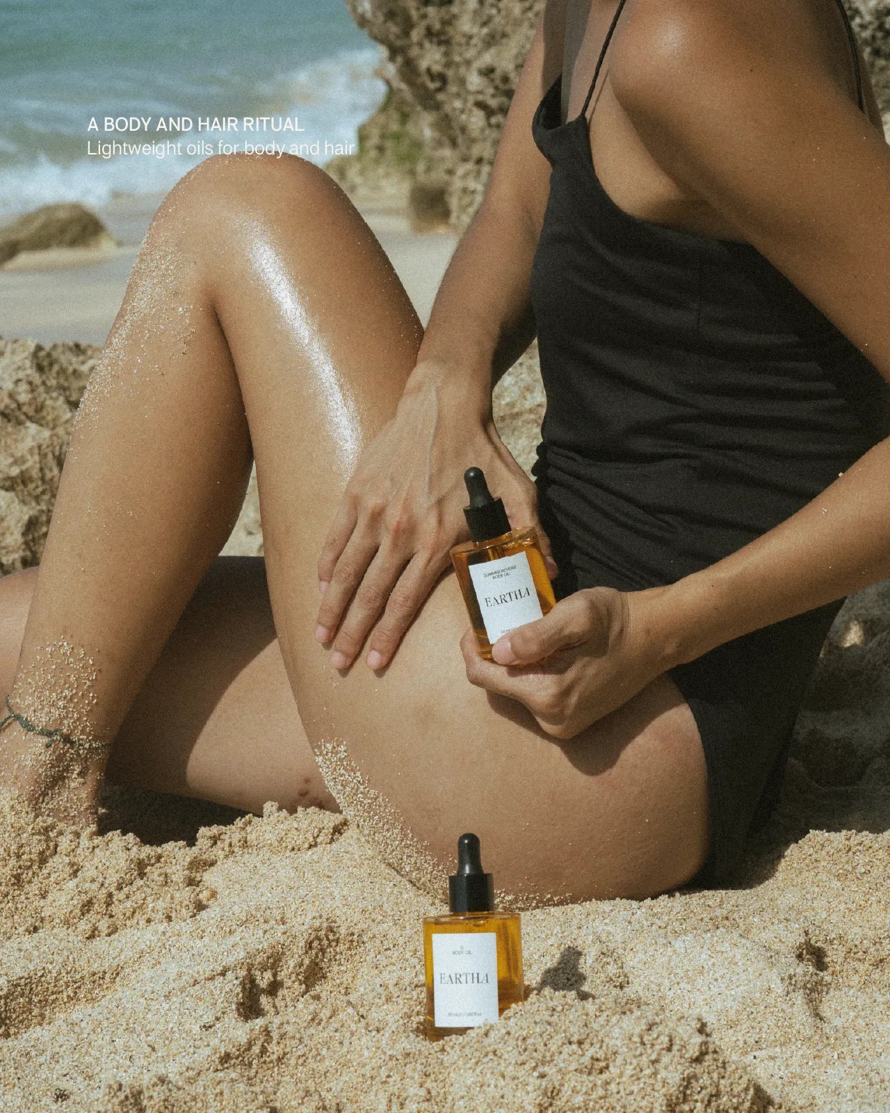 Person on a beach holding a bottle of 'Eartha' product with another bottle on the sand.