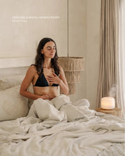 Woman practicing breathwork on a bed in a bedroom with a diffuser lamp next to her.